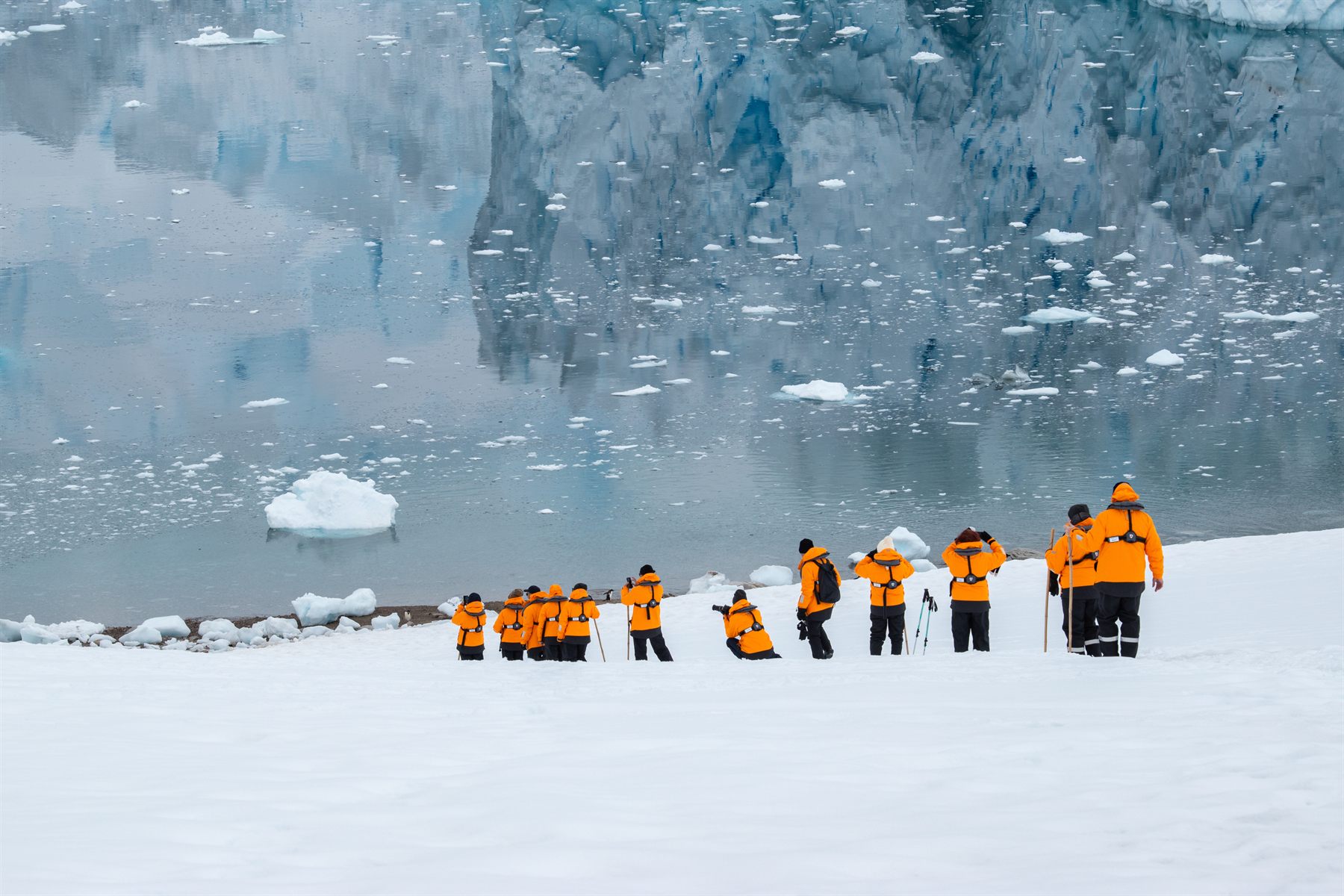 Expedition guests in orange parkas on the snow below an Antarctic glacier front